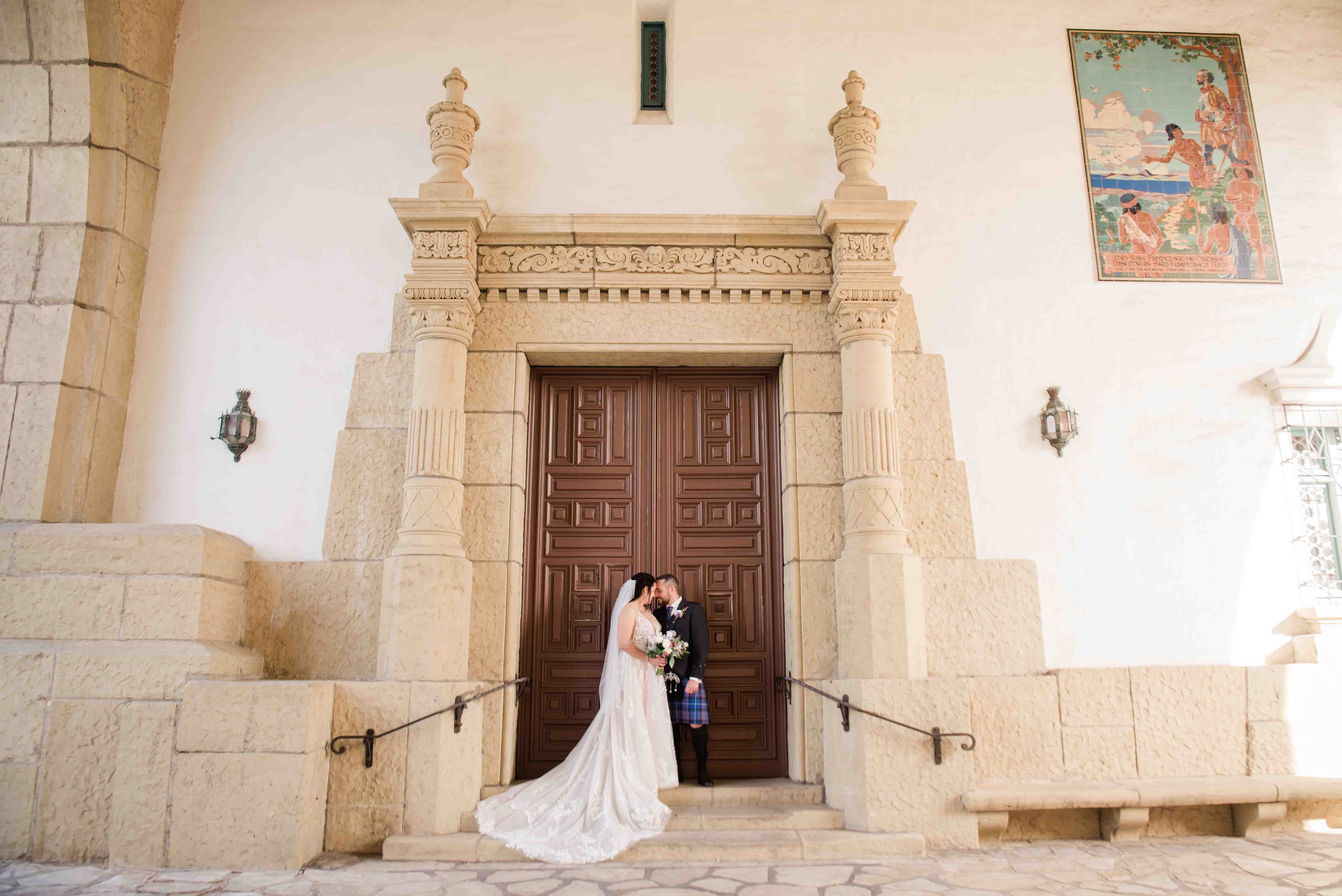 groom ins Scottish kilt forehead to forehead to bride in front of large decorative door at santa barbara courthouse