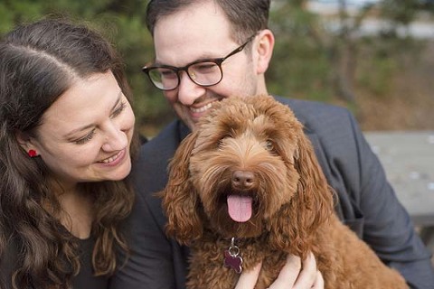 caucasian couple sitting next to each other. He looks at his fiancé while she looks at their dog on his lap