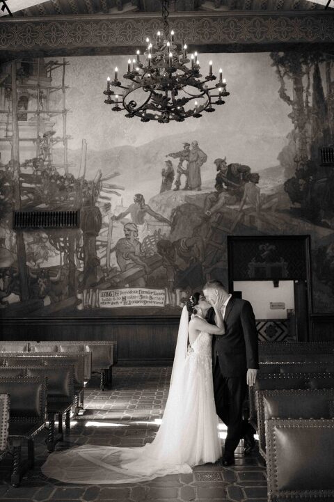 bride and groom kissing under chandelier in the mural room of santa barbara courthouse