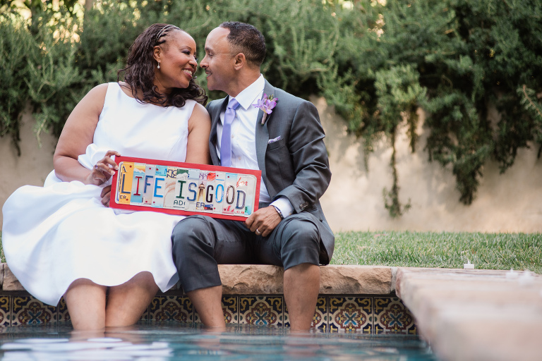 Black bride and groom in Santa Barbara California sitting in pool rented from Air BnB holding a sign saying "Life is Good."