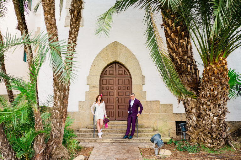 bride and groom in white and purple outfits standing in from on decorative door under palm trees at santa Barbara courthouse