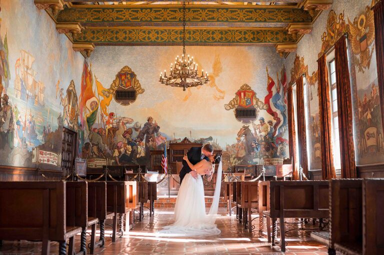 bride and groom kissing under chandelier in the mural room of santa barbara courthouse