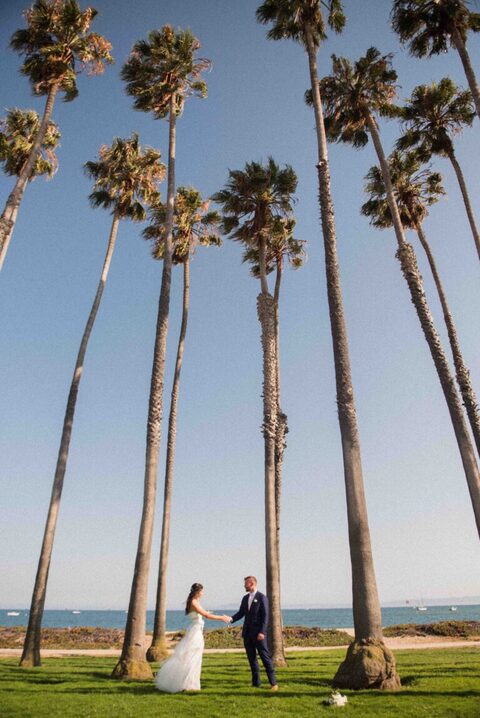 bride and groom holding hands under palm trees at east beach with pacific ocean in background
