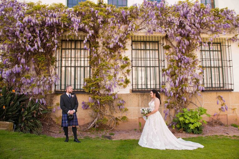 groom in Scottish kilt looking at his bride under a frame of purple wisteria at santa Barbara courthouse